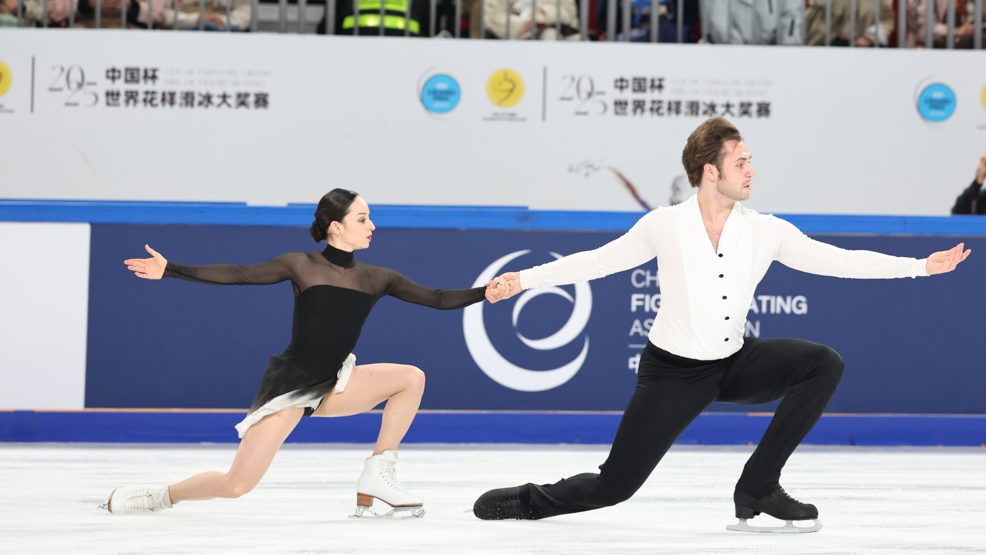 Katie McBeath and Daniil Parkman kneel in unison while holding hands. Katie (left) is a young woman with black hair tied back in a low bum wearing a black skating costume with sheer long sleeves. Daniil (right) is a man with short brown hair wearing a long sleeve white button down top and black pants