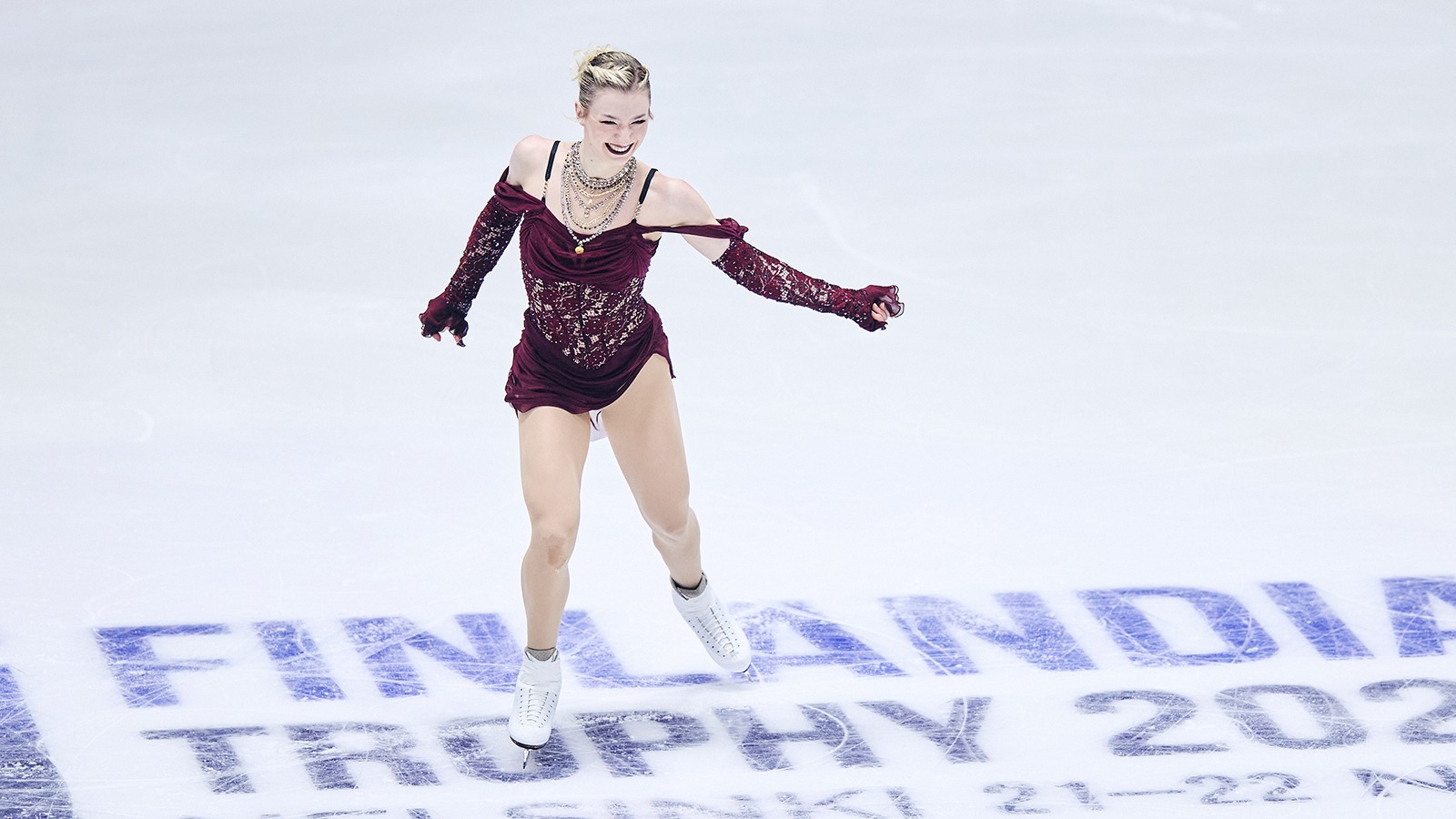 Amber Glenn, in a maroon dress with cold shoulder sleeves and her hair up, skates at center ice at 2025 Finlandia Trophy. She smiles as she skates.