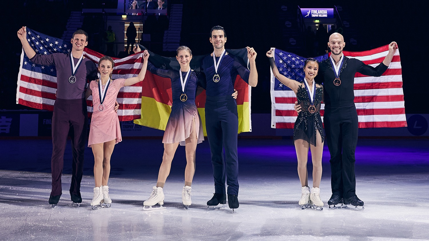 The 3 pairs teams who medaled at Finlandia Trophy. To the left, Alisa Efimova and Misha Mitrofanov, in the middle the German team and to the right Ellie Kam and Danny O'Shea. The two U.S. pairs teams stand holding U.S. flags behind them, the Germans hold their flag behind them. All smile with medals around their necks.