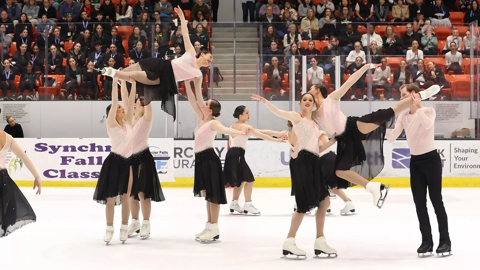 The Haydenettes perform a lift with two skaters hoisted in the air. They are all wearing matching skating costumes with a light pink top and black skirt. One male skater is wearing a coordinating top with black pants.