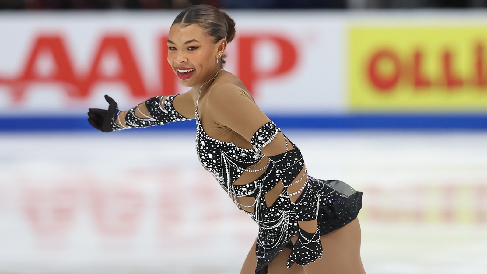 Starr Andrews skates with a smile on her face and her right arm outstretched. Starr is a young Black woman with her hair tied back in a bun wearing a black skating costume with white jeweled details. 