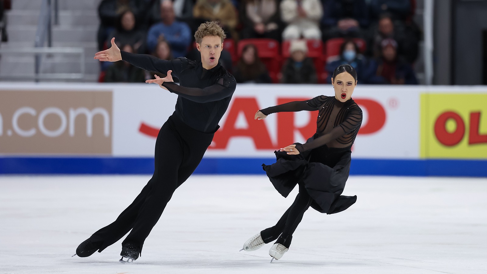 Madison Chock and Evan Bates skate side by side with both athletes pointing their arms to the left in unison. Evan (left) is a tall man with short blonde hair wearing a long sleeve black top and black pants. Madison (right) is a woman with black hair tied back in a bun. She is wearing a black skating costume with long sleeves and a long black skirt