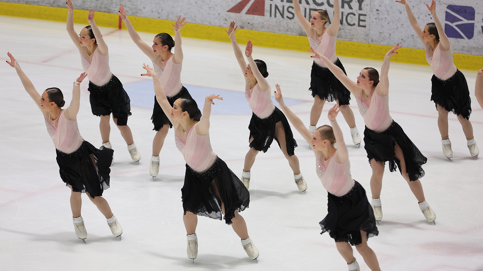 The Haydenettes skate in unison in a group with their hands raised above their heads. They are all wearing matching costumes with a long sleeve pink top and a black skirt.