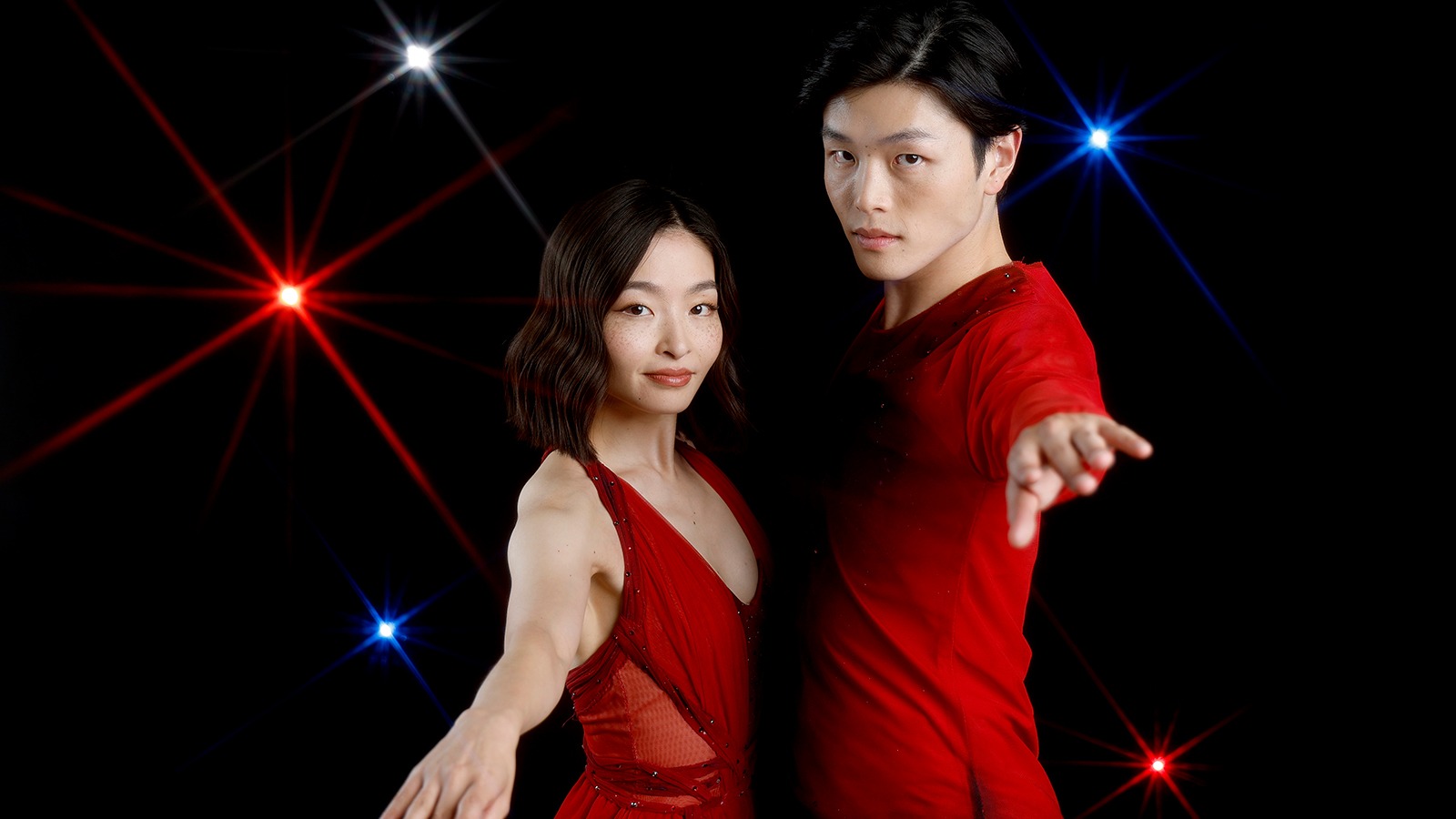 Maia Shibutani (left) and Alex Shibutani (right) stand in front of a black background with red, white and blue lights facing each other with their arms extended. Maia is a young woman with short shoulder length black hair wearing a sleeveless red skating costume. Alex is a young man with short black hair wearing a matching red long sleeve top. 
