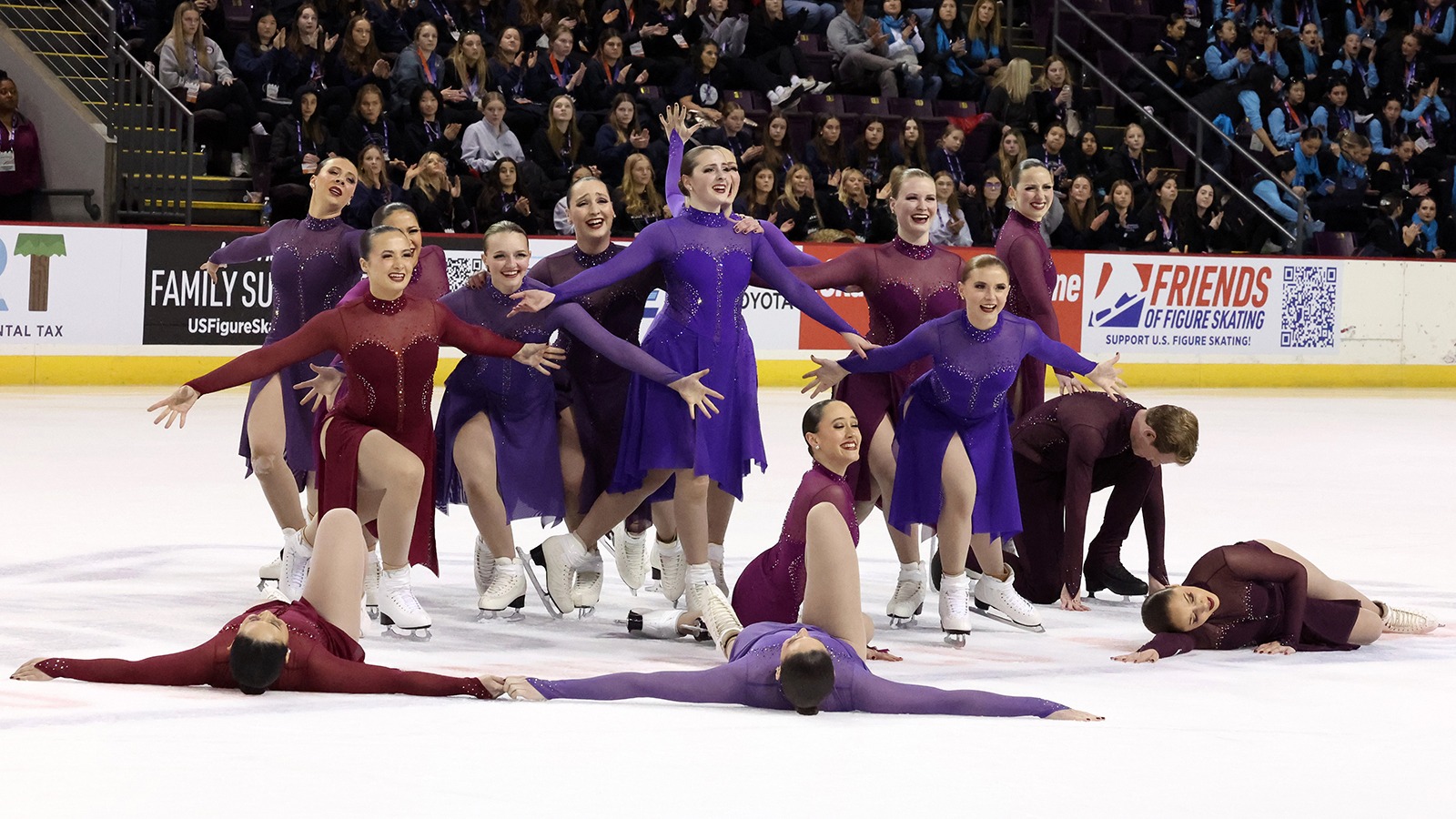 The Haydenettes strike their ending pose with smiles on their face. The team is wearing coordinating purple or red long sleeve skating costumes.