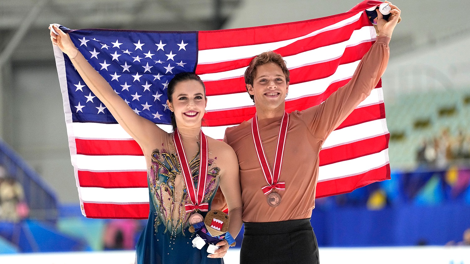 Caroline Green (left) and Michael Parsons (right) hold up the U.S. flag behind them and smile during the medal ceremony at NHK Trophy. They have bronze medals around their neck and they both smile.
