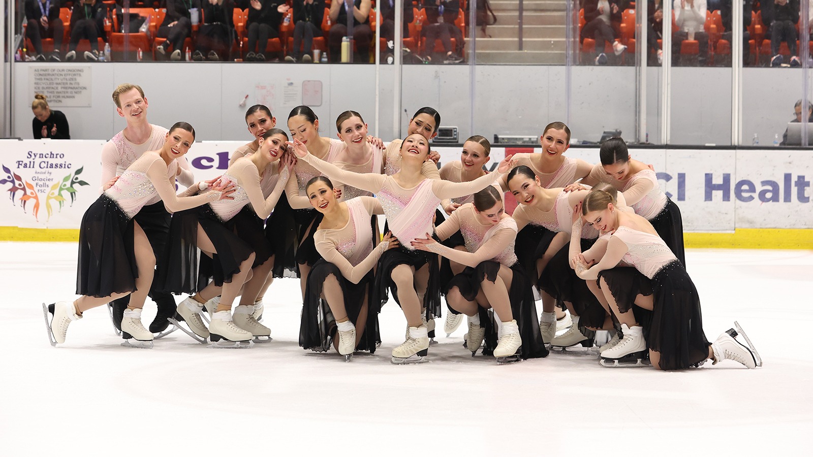 The Haydenettes strike a beginning pose, leaning on each other. The skater in the middle looks up with a smile on her face and her arms outstretched. They are all wearing matching skating costumes with a light pink long sleeve top and black skirt. One male skater on the far left is wearing a matching pink top with black pants.