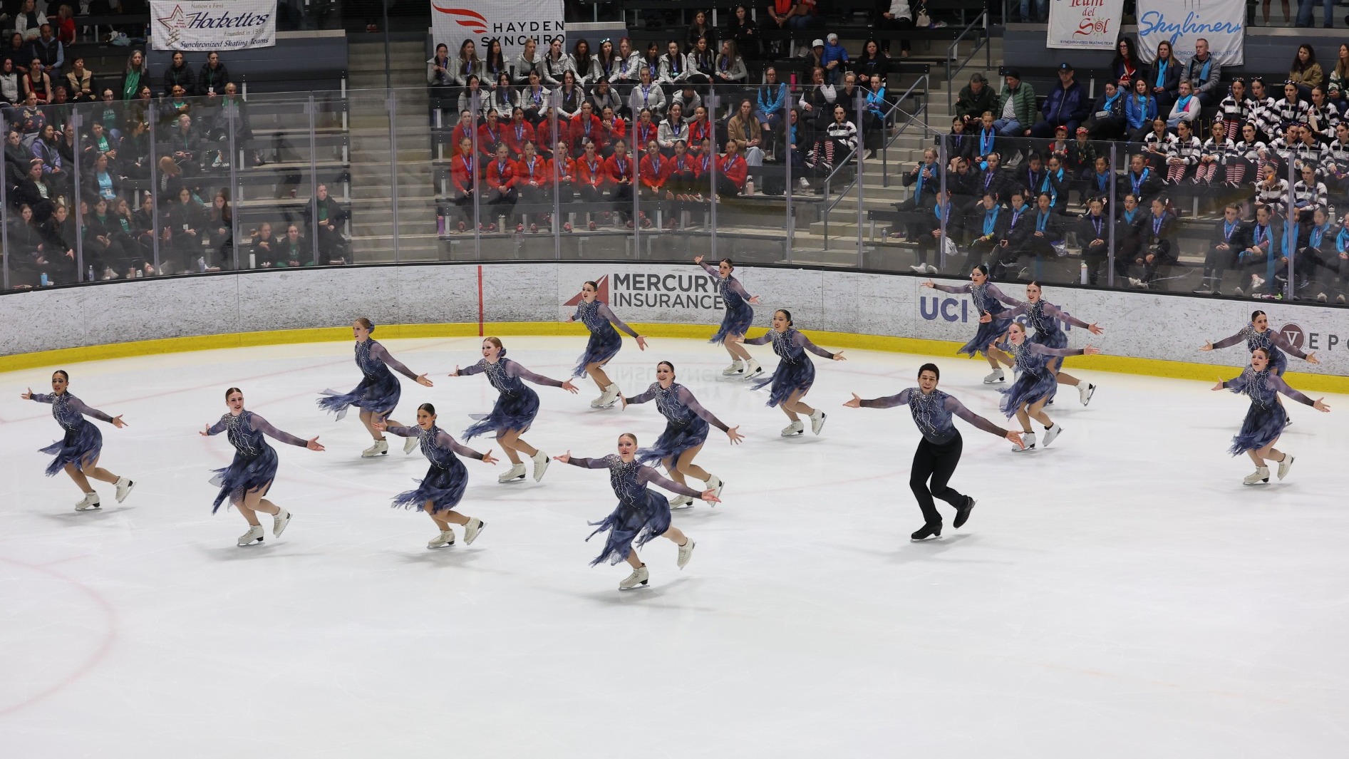 Taken from above, Teams Elite, in dark blue/grey costumes, skates as a group to the left, their arms out to their sides.