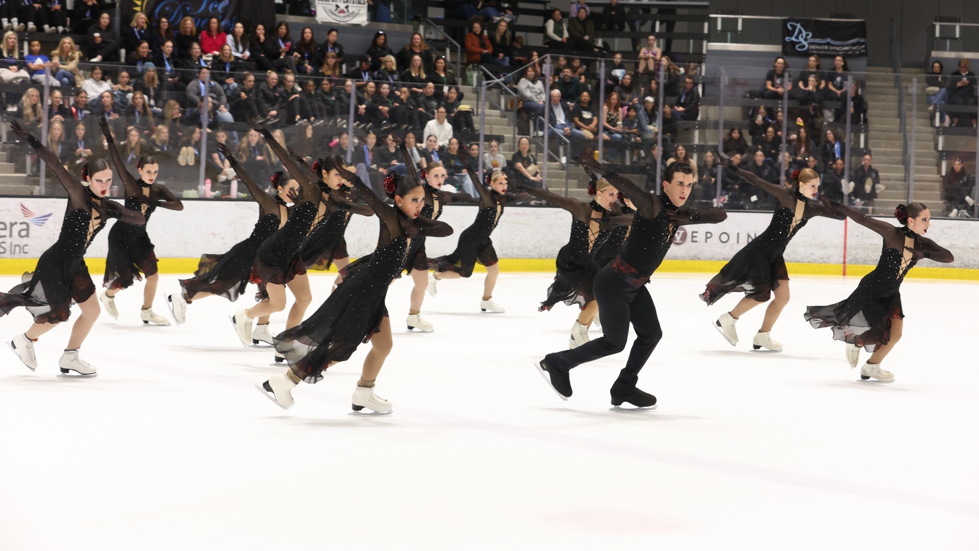 The Teams Elite junior synchro team skates in unison with both arms pointing to the left. They are all wearing matching black skating costumes with long sheer sleeves, a long black skirt and a red flower in their hair. One male skaters in the front row is wearing a matching top with black pants and a red belt.  