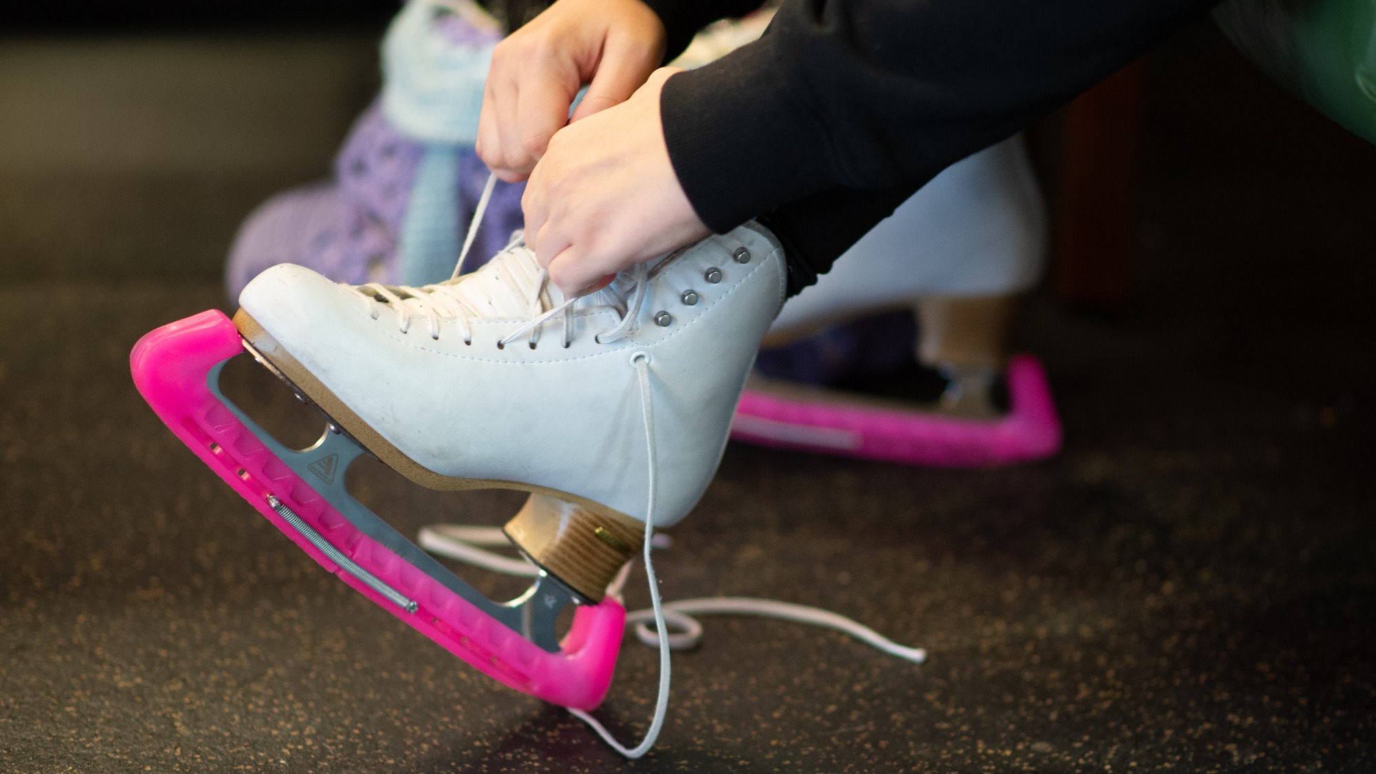 close up of a girl tying up her white figure skates with pink skate guards on them. 
