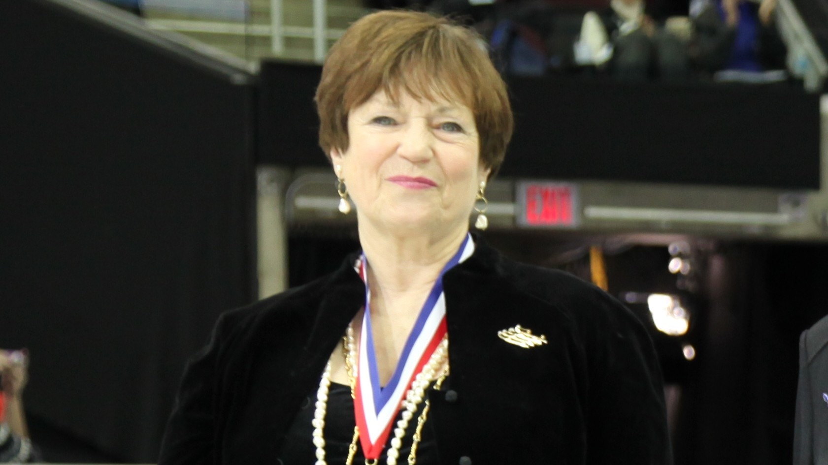 Photo of Sonya Dunfield at her induction ceremony to the U.S. Figure Skating Hall of Fame. Sonya is an older woman with short brown hair wearing a black top with black jacket over top and a medal with a red, white and blue ribbon. She is wearing a gold necklace, gold earrings and a gold brooch
