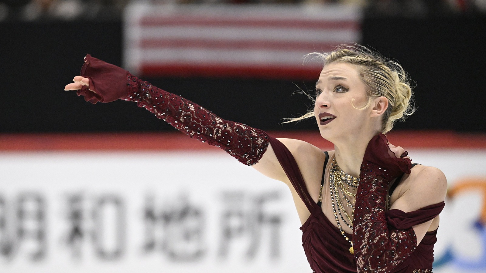 Photo taken from the waist up of Amber Glenn. She looks off to the left with one arm outstretched. Amber is a young woman with blonde hair tied back in a bun wearing a maroon skating costume with chunky gold necklaces and matching maroon gloves.