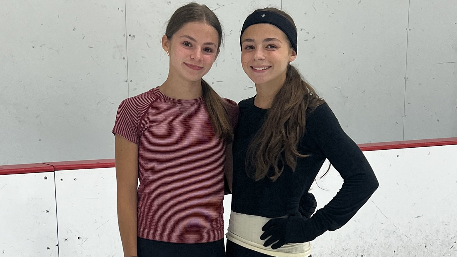 Quinn Cushing, wearing a red T-shirt, stands next to twin Harlow Cushing, in black, near the rink boards of an ice rink.
