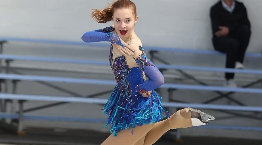 Wearing a blue costume, mouth open, Isobel Carney shows her presentation skills with one arm bent below her face and the other on her waist, gliding on one leg. 