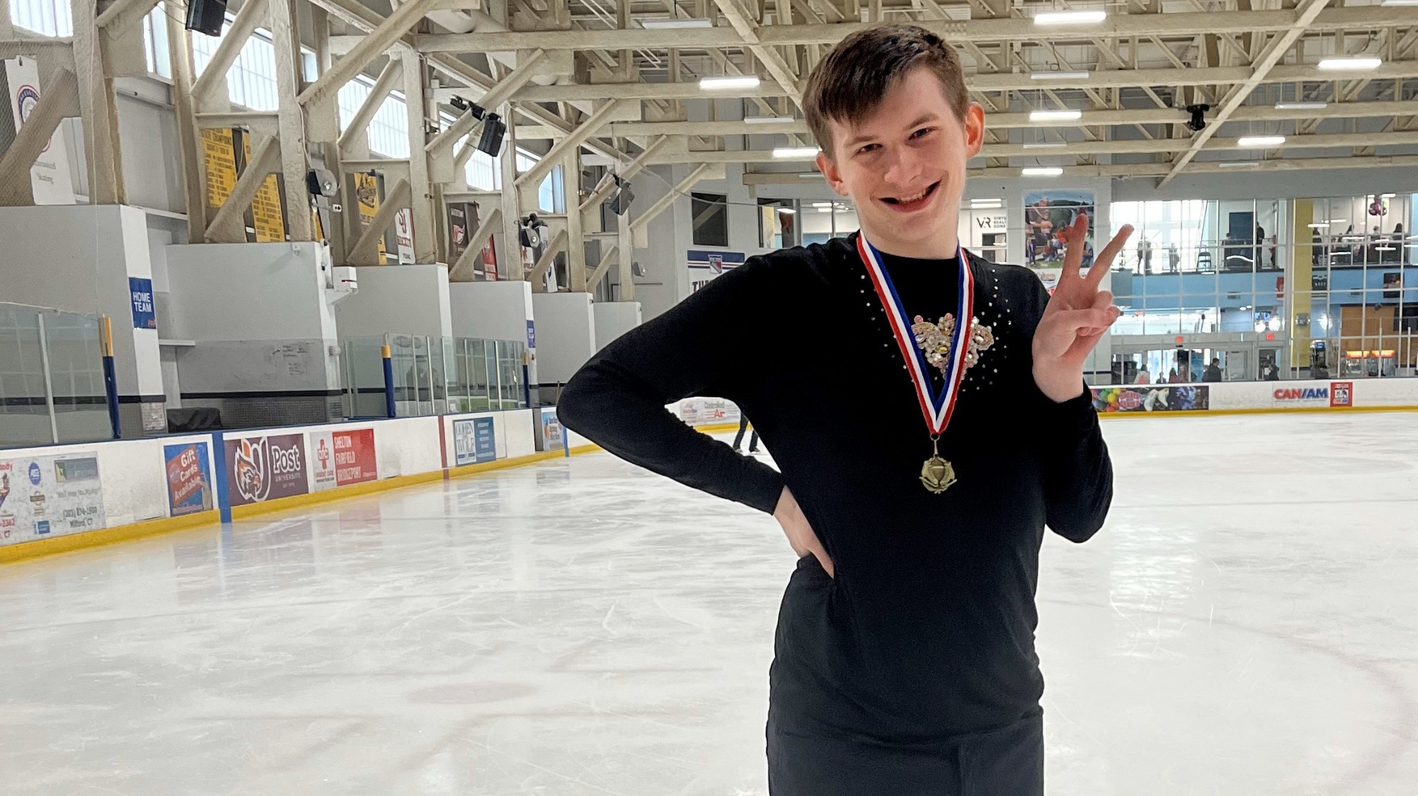 Tim poses on the ice with a medal around his neck. Tim is a white teenager with short brown hair. He is wearing a black long sleeved shirt with jewels on the chest. He stands with his right hand on his hip and his left hand making a peace sign