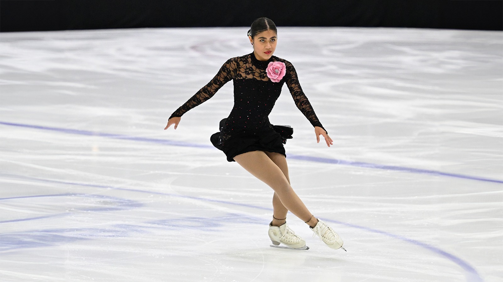 Amal Israilova skates with one leg out in front of her body. She is a young woman with black hair tied back in a bun wearing a long sleeve black skating costume with a large pink flower on the lapel