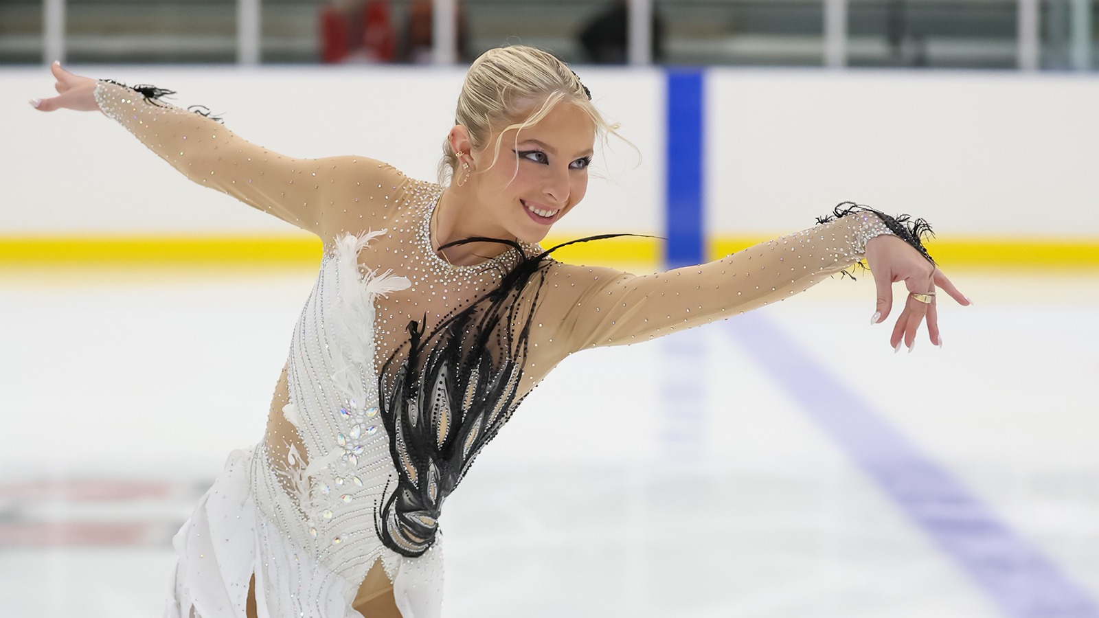 In a photo taken from the waist up, Brooke Tufts skates with her arms outstretched, looking to the right. She is a young woman with blonde hair tied back in a bun wearing a white skating costume with black feathers inspired by Swan Lake. 