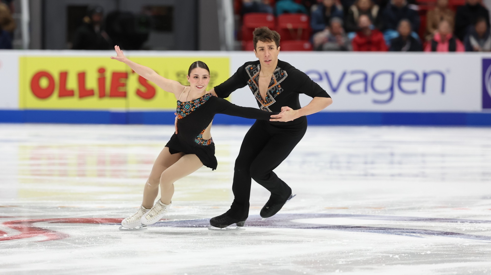 Valentina Plazas and Maximiliano Fernandez skate together with Valentina in front with her arms outstretched and Maximiliano close behind with one hand on her waist and the other holding her left hand. Valentina is a Latina woman wearing a long sleeve black skating costume with colorful geometric details. Maximiliano is a Latino man wearing a long sleeve black top with a deep v-neck and matching geometric details along the collar. 
