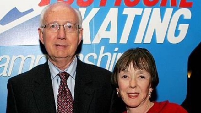 In a suit with red tie, Ron Hershberger stands next to his wife Gayle, in front of a U.S. Figure Skating Championships sign.