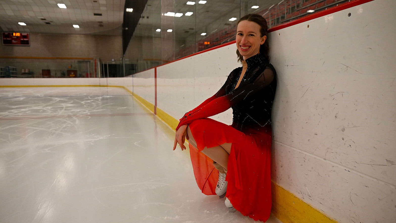 Alexandra Poret, in a black top and pink dress, kneels down rinkside with a smile.