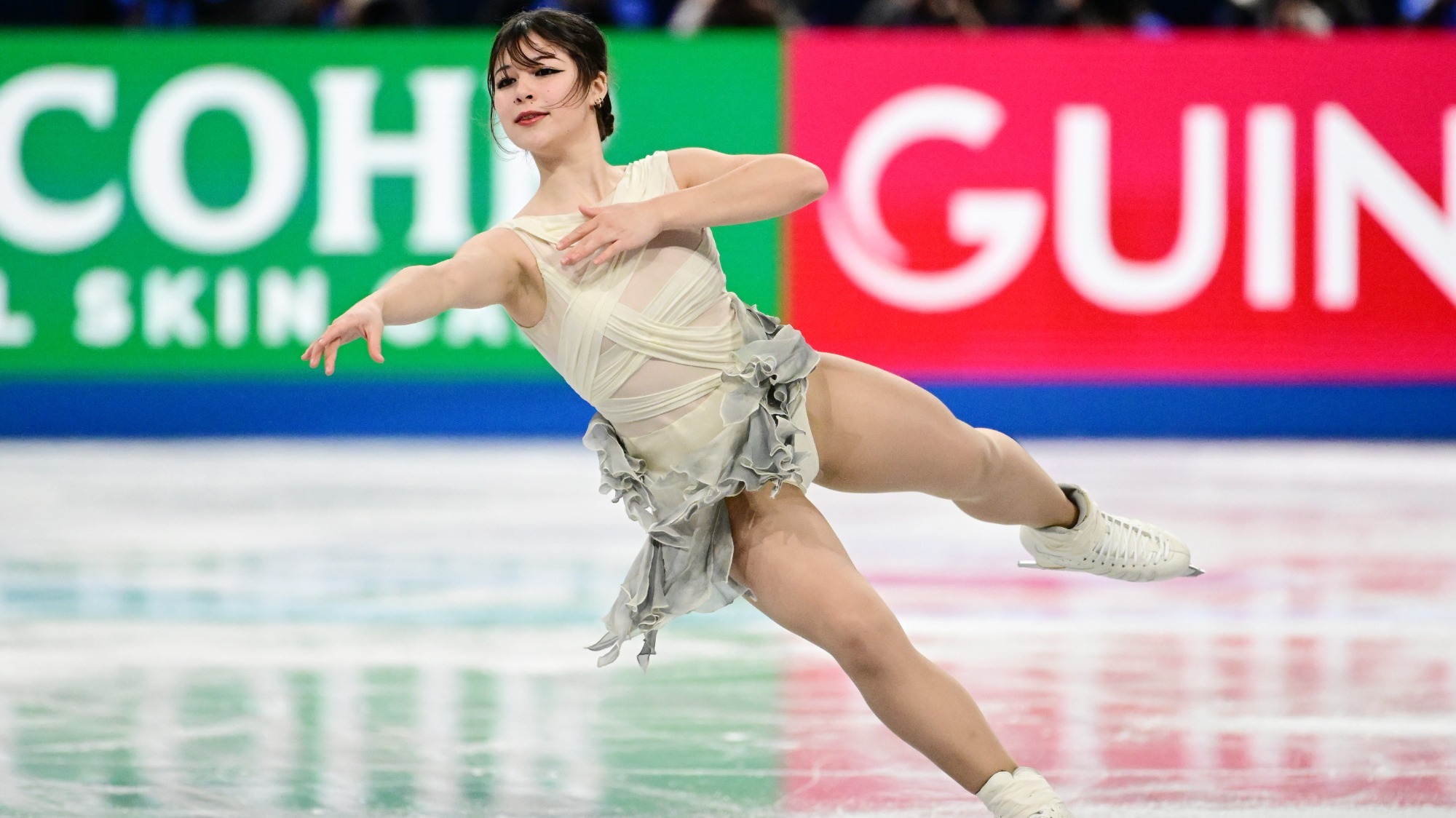 Alysa Liu skates on one leg with the other extended behind her. She holds one arm extended outward and the other close to her chest. Alysa is a young woman with brown hair tied back in a low bun. She is wearing a white skating costume with the fabric draping over her body