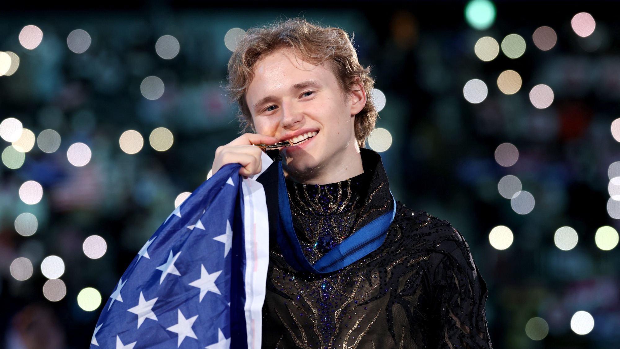Photo taken from the waist up of Ilia Malinin biting his Grand Prix Final gold medal with an American flag draped around his shoulders. Ilia is a young man with blonde hair wearing a long sleeve black top with dark blue and gold details down the front