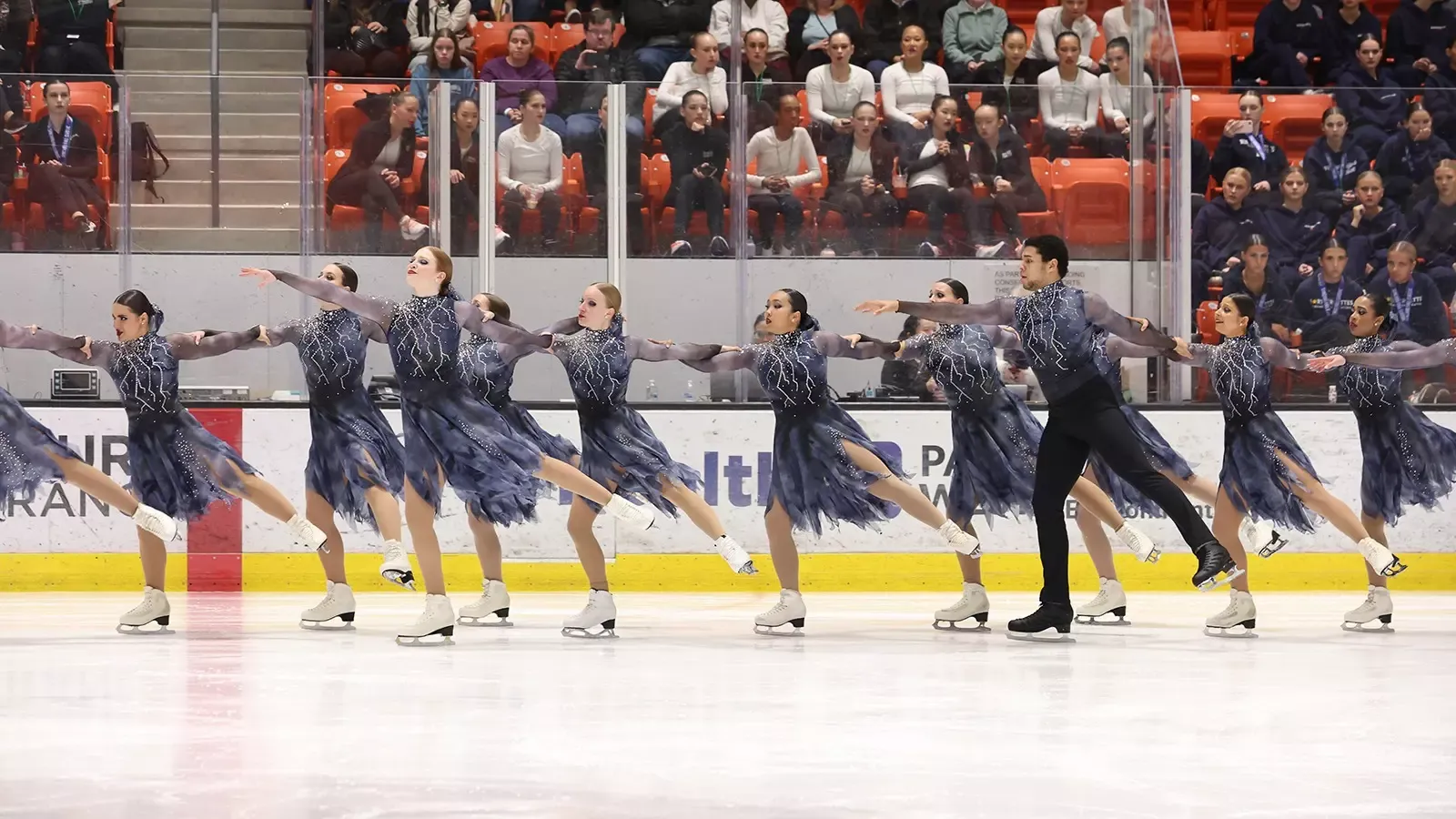 Teams Elite senior, dressed in gray, glide across the ice in a two-line formation.