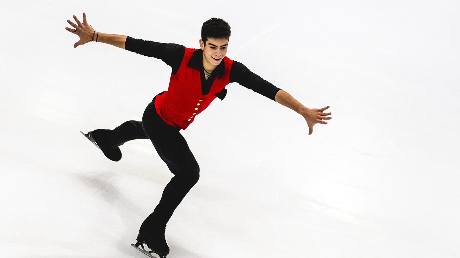 Dressed in red with black sleeves and black pants, Jacob Sanchez glides on the ice, arms fully extended to his sides.