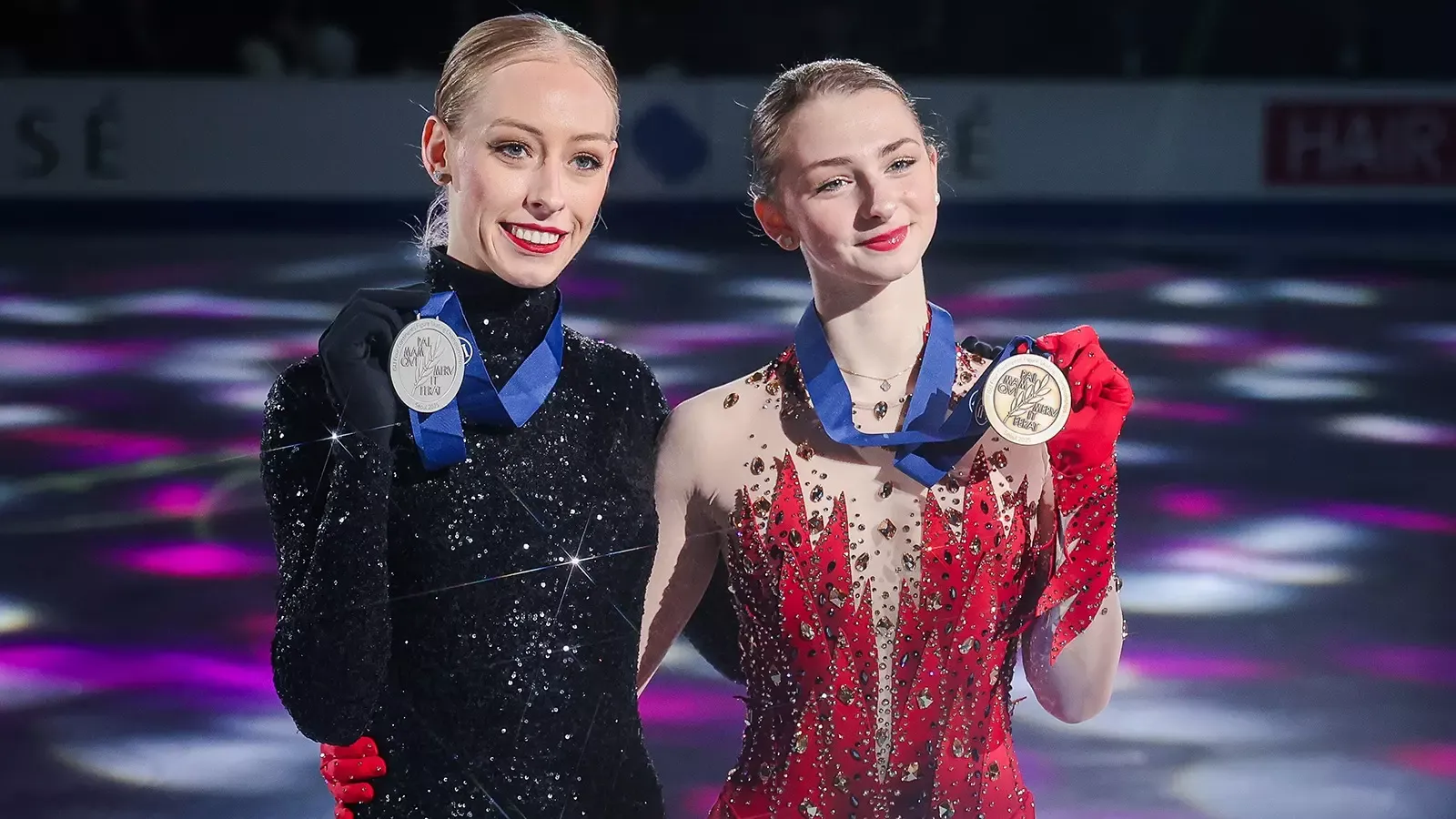 Bradie Tennell and Sarah Everhardt pose with their Four Continents medals, smiling at the camera with their arms around each others waist. Bradie (left) is a young woman with blonde hair tied back in a bun wearing a long sleeve black and sparkly skating costume. Sarah (right) is a young woman with blonde hair tied back in braids wearing a red skating costume designed to resemble feathers with matching red gloves. 