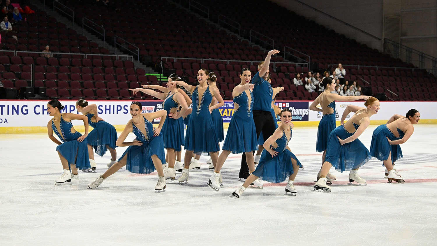 Esprit de Corps, an adult team, is wearing blue dresses. They hit their starting pose at center ice of the 2024 U.S. Synchronized Skating Championships. With skaters in a circle, the women in the center stand while the ones on the outside are crouched. All are hitting a pose and smiling. 