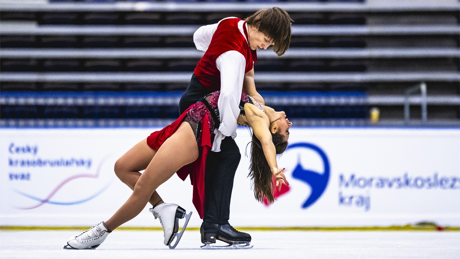 Dimitry Tsarevski dips Katarina Wolfkostin during their performance on ice. Katarina is a young woman with long brown hair wearing a sleeveless red skating costume. Dimitry is a young man with shirt brown hair wearing a long sleeve white shirt with a red vest over top and black pants. 