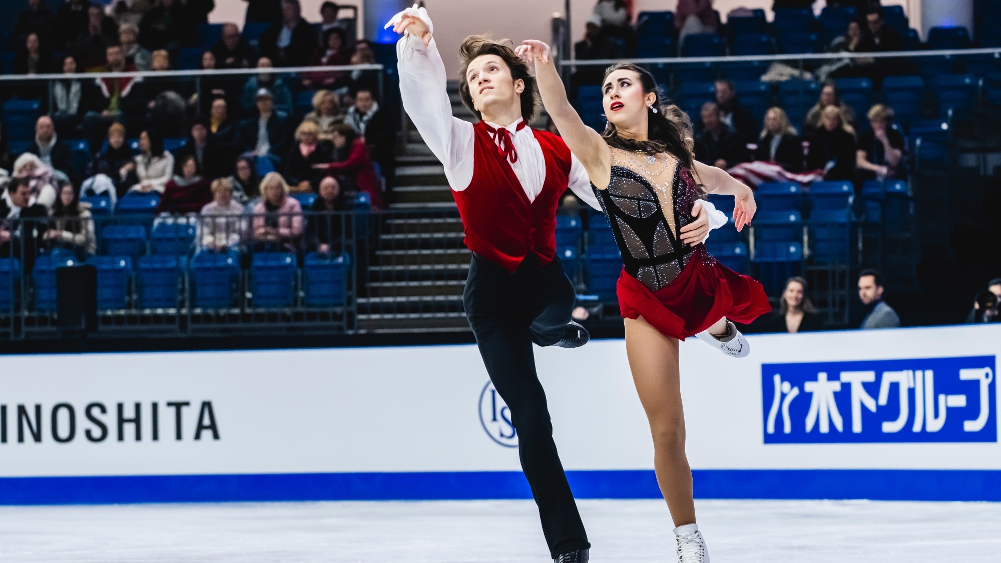 Katarina Wolfkostin and Dimitry Tsarevski skate to the left with one arm extended outward and one leg raised behind them. Dimitry (left) is a man with shirt brown hair wearing a white button down shirt with a red vest on top and black pants. Katarina is a young woman with long brown hair wearing a  skating costume with a black corset top and red skirt.