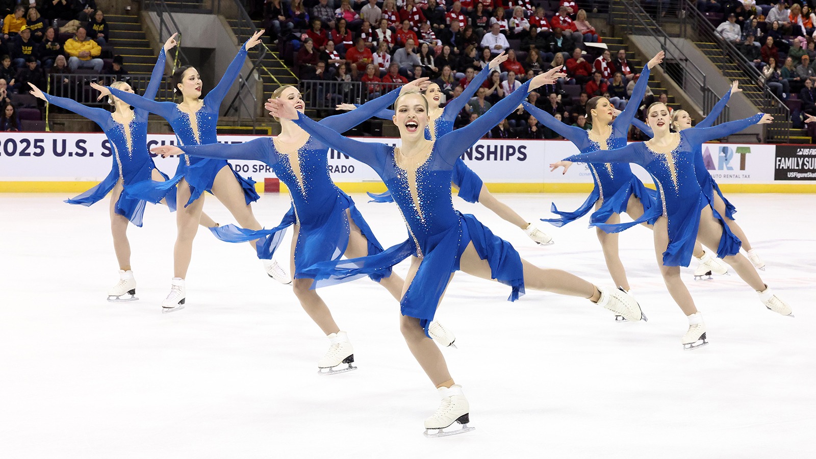 The Haydenettes skate in unison with their arms outstretched and one leg extended behind them. They are all wearing matching long sleeve blue skating costumes with a deep n neck and jewel detailing around the neckline. They all have smiles on their face. 