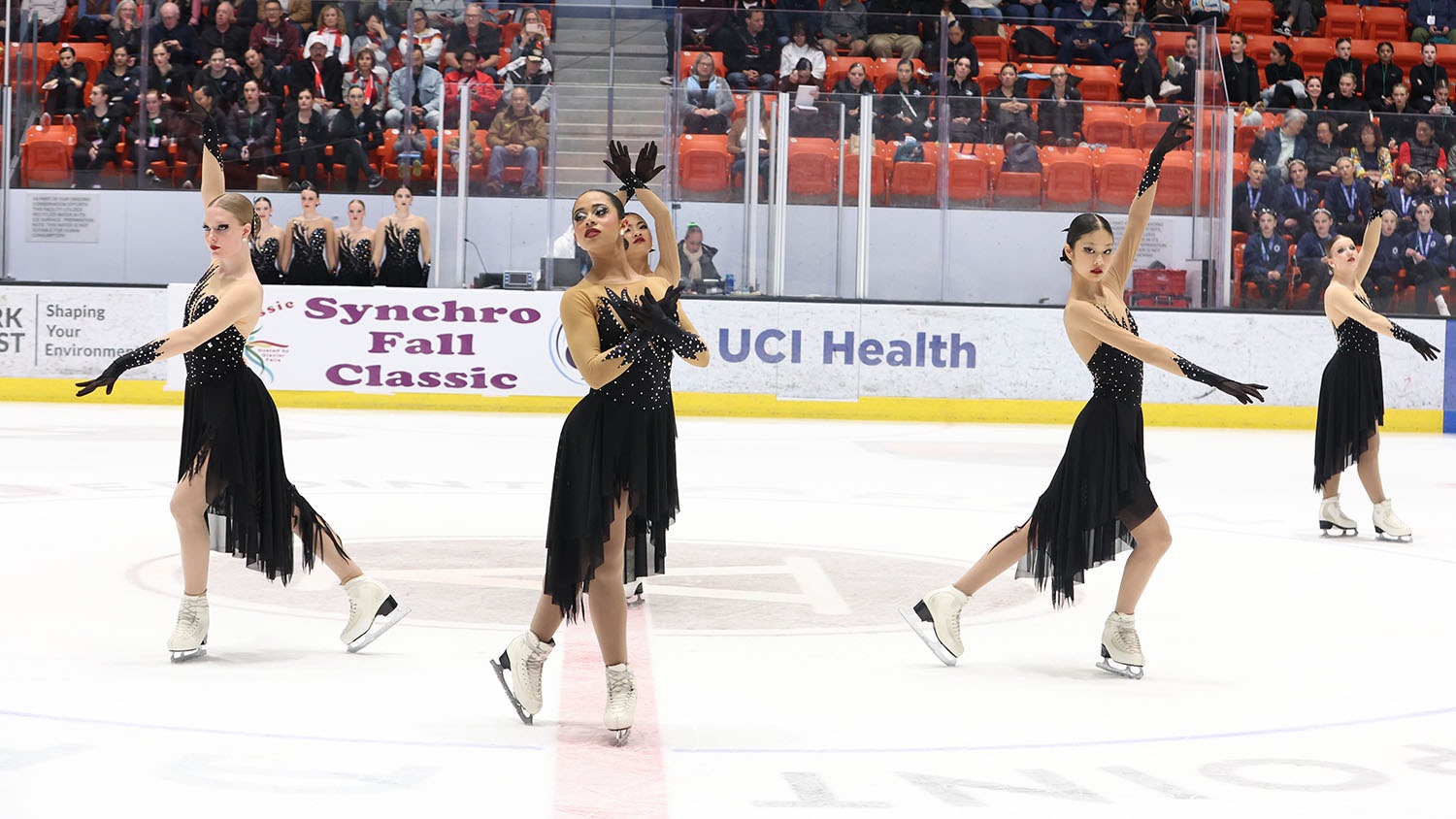 Four Teams Elite skaters in their opening pose on center ice. All wear black dresses. The two skaters on the right face right while the skater on the left mirrors their pose. The skater in the center holds her arms across her chest.