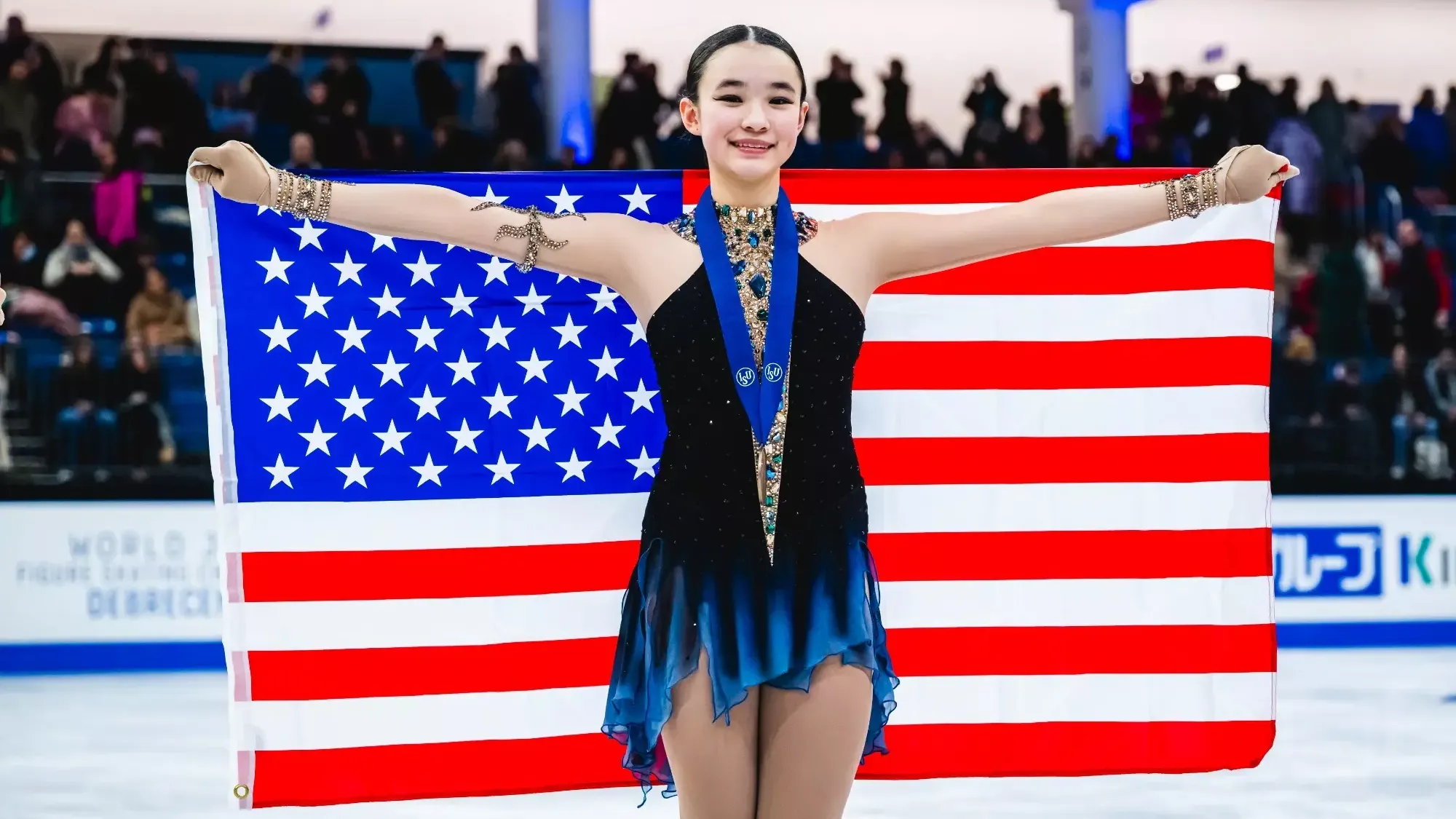 Elyce Lin-Gracey stands holding an American Flag behind her with her bronze medal around her neck, Elyce is a young Asian woman wearing a black skating costume with blue along the bottom. and jewel detailing along the front. 