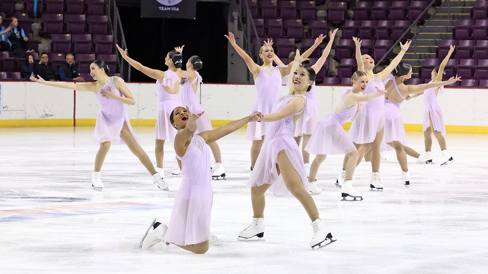 The Hockettes perform wearing coordinating light purple skating costumes. All athletes are standing except one who is on their knees. They all have smiles on their face and their arms raised in the air