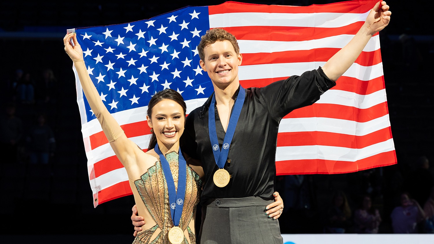 Madison Chock and Evan Bates celebrate their gold at the victory ceremony of the 2025 World Championships. They hold the U.S. flag behind them, gold medals around their necks.