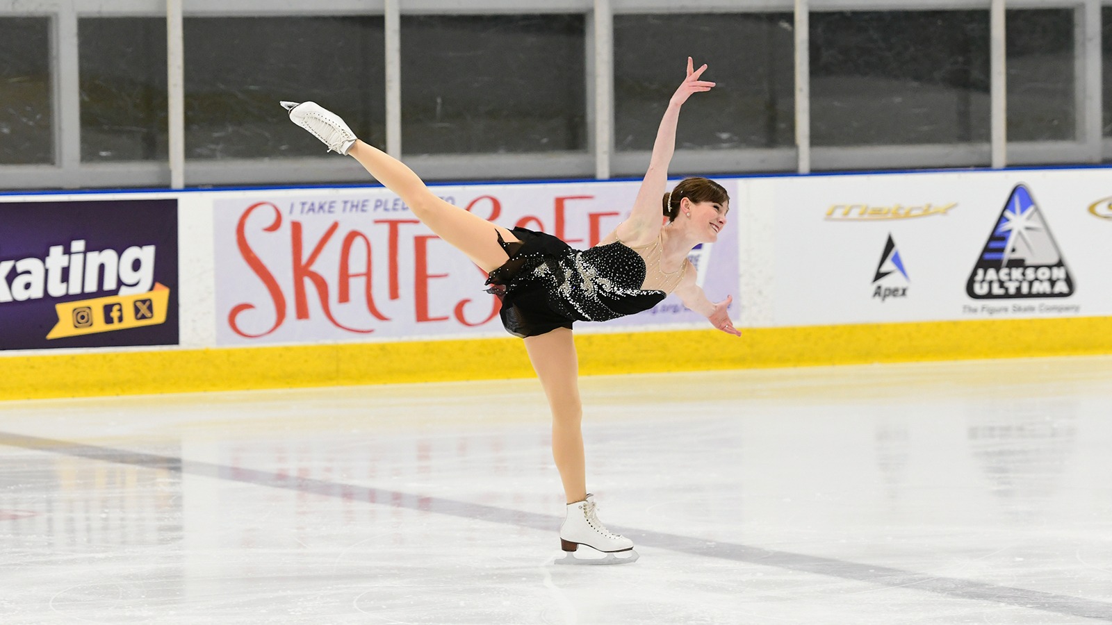 Kristina Struthwolf-Leato performs a spiral leaning forward with her arms outstretched and one leg extended. She is a woman with brown hair tied back in a low bun. She is wearing a black skating costume with silver jewel details