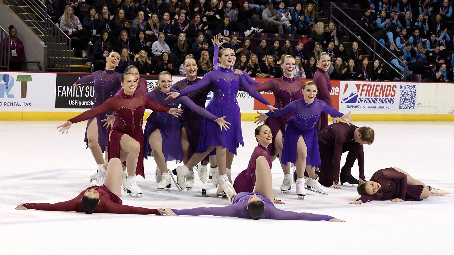 The Haydenettes, in dresses of different shades of purple, hit their ending pose of their free skate. Three athletes lay on the ground with others kneeling and standing behind them. They are smiling.