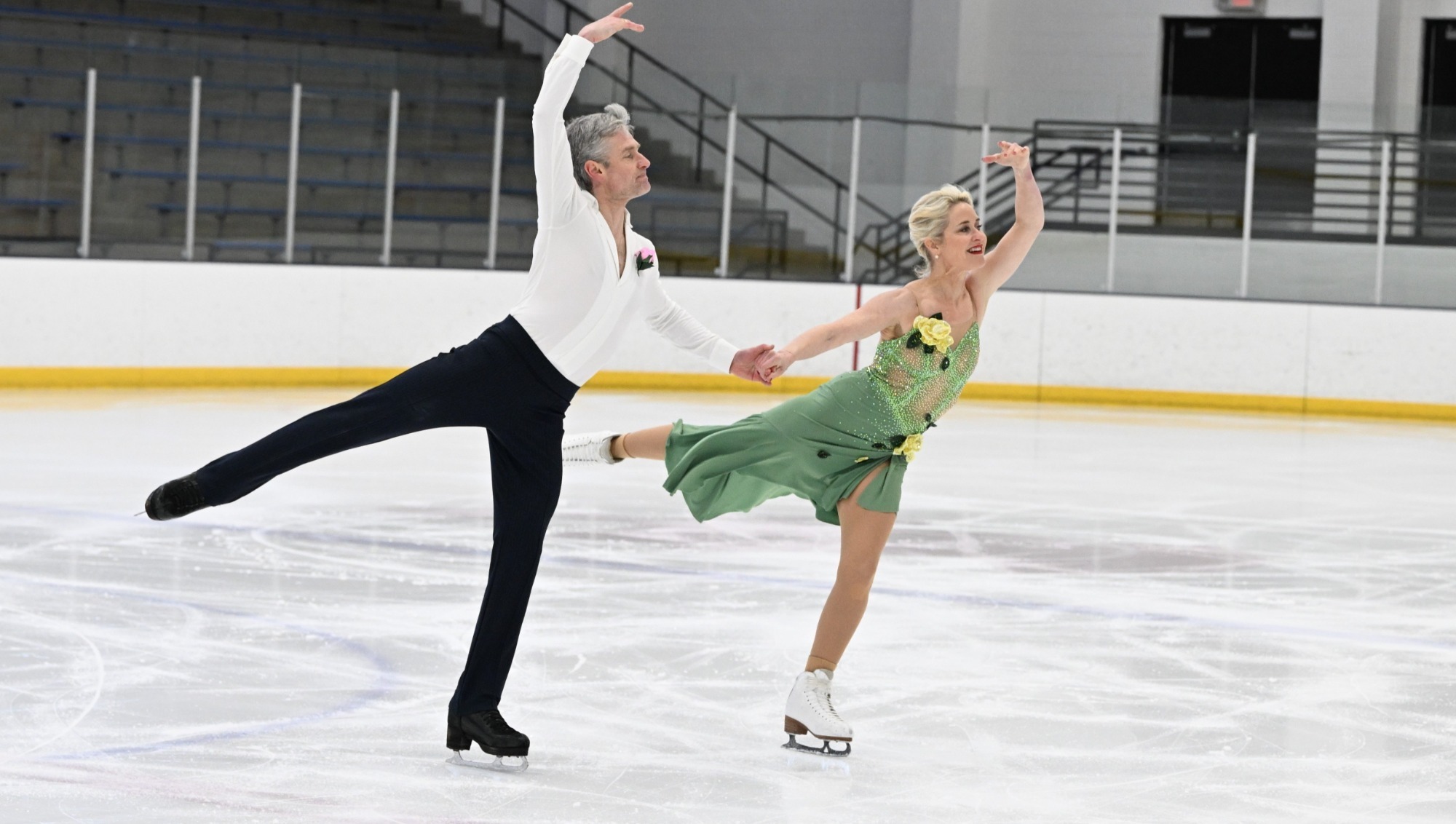 Jennifer Freedman and Oleg Voyko compete together on ice. Oleg (left) is an older man with short grey hair wearing a white button down shirt and black pants. Jennifer (right) is an older woman with blonde hair wearing a green and yellow skating costume with yellow flowers on the front. They skate hand in hand. 
