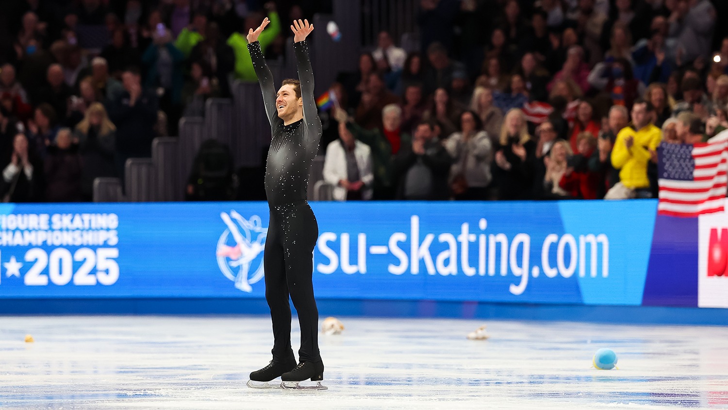 Jason Brown stands at center ice and salutes the crowd at the World Championships. His arms are both up as he waves, a smile on his face. He is wearing black skates, pants and a grey to black ombre long sleeve shirt. The crowd gives him a standing ovation in the background.