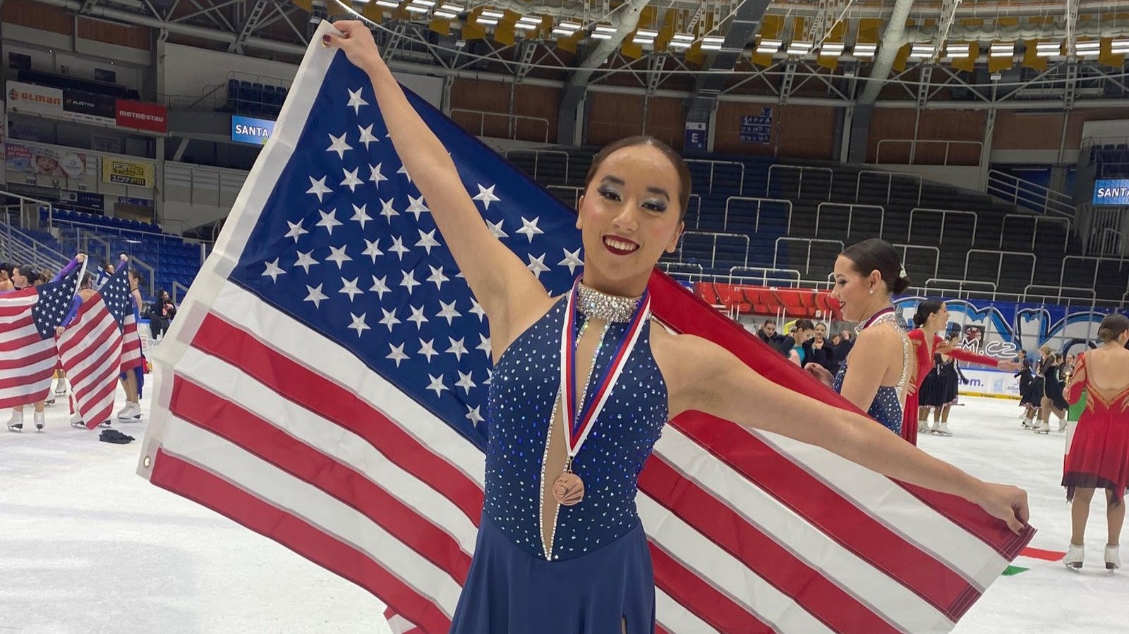 Evelyn Duffy poses holding an American flag with a medal arounf her neck. Evelyn is a young Asian American woman with her hair tied back in a bun wearing a dark blue skating costume