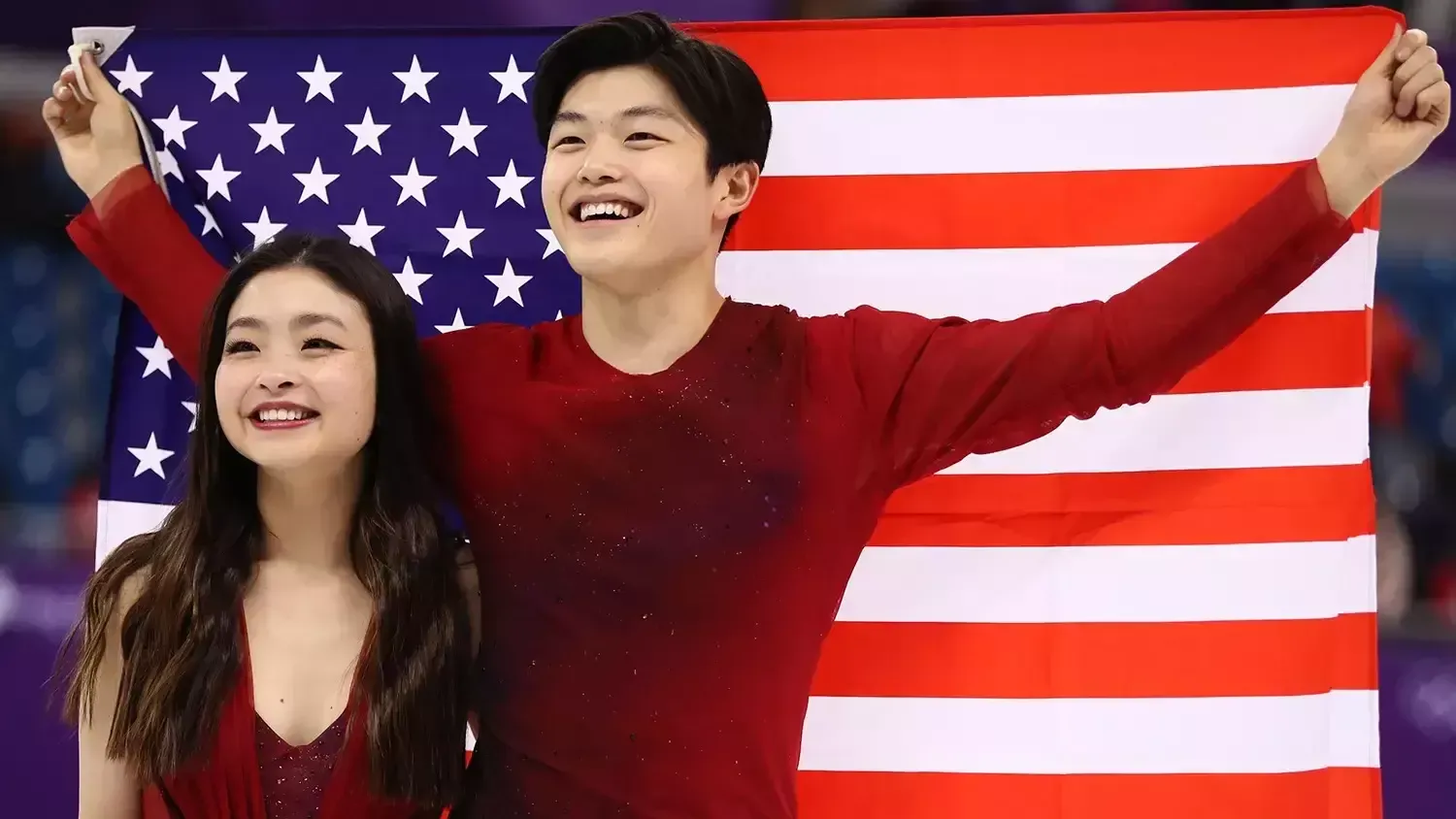 Maia and Alex Shibutani, both wearing red, smile as they look upwards during the victory ceremony at the 2018 Olympics. Alex holds a USA flag behind them. The photo is taken from the waist up.