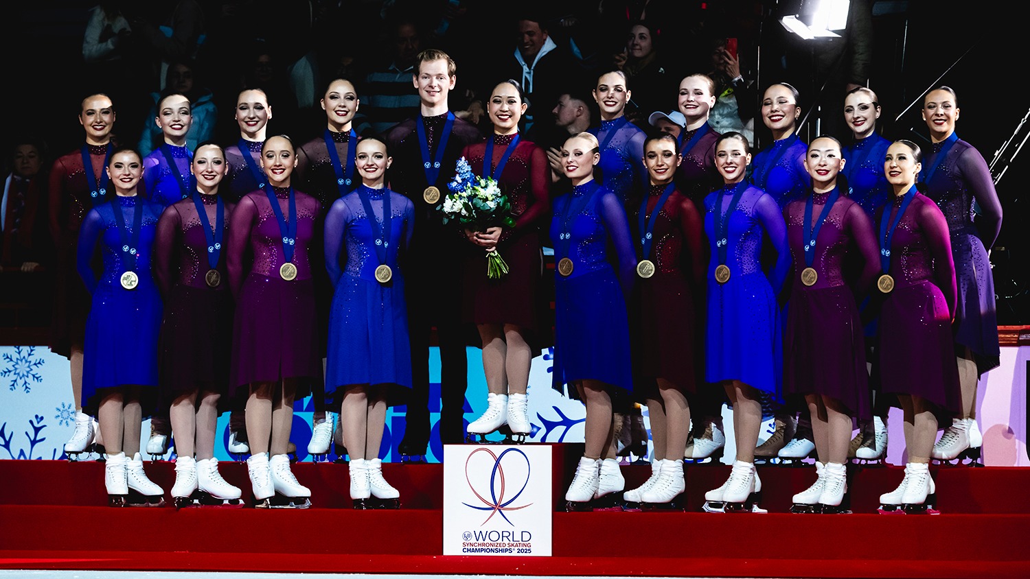 The Haydenettes, wearing their free skate dresses which are shades of purple, stand on the podium at the World Synchronized Skating Championships, bronze medals around their necks. They all smile.