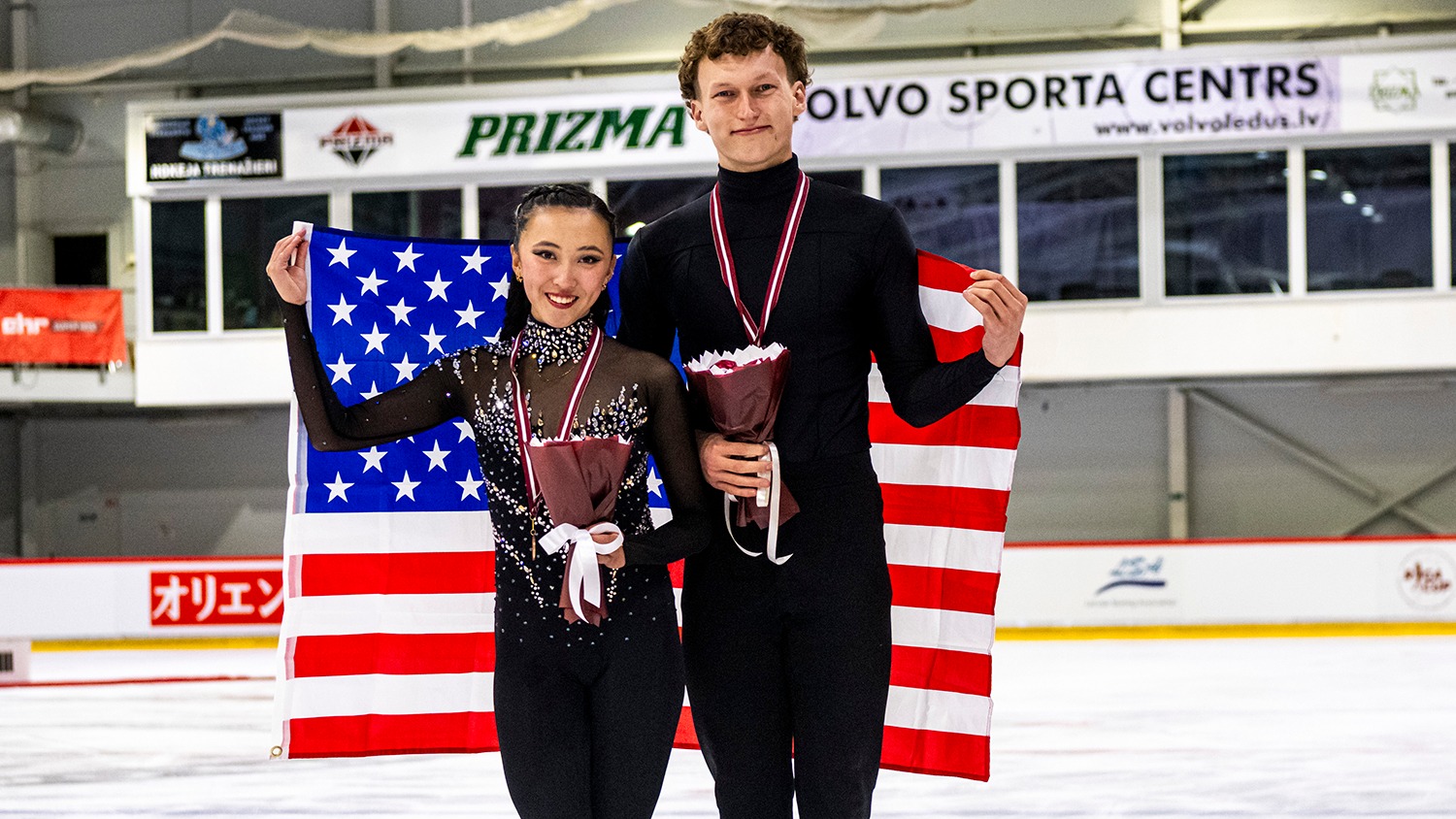 RIGA, LATVIA - AUGUST 22: Bronze medal winners Naomi Williams and Lachlan Lewer of the United States pose with their medals during medal ceremony after the ISU Junior Grand Prix of Figure Skating at Volvo Sporta Centrs on August 22, 2025 in Riga, Latvia. They stand on the ice, holding the U.S. flag behind them and holding a bouquet of flowers. Both wear black skating costumes.
