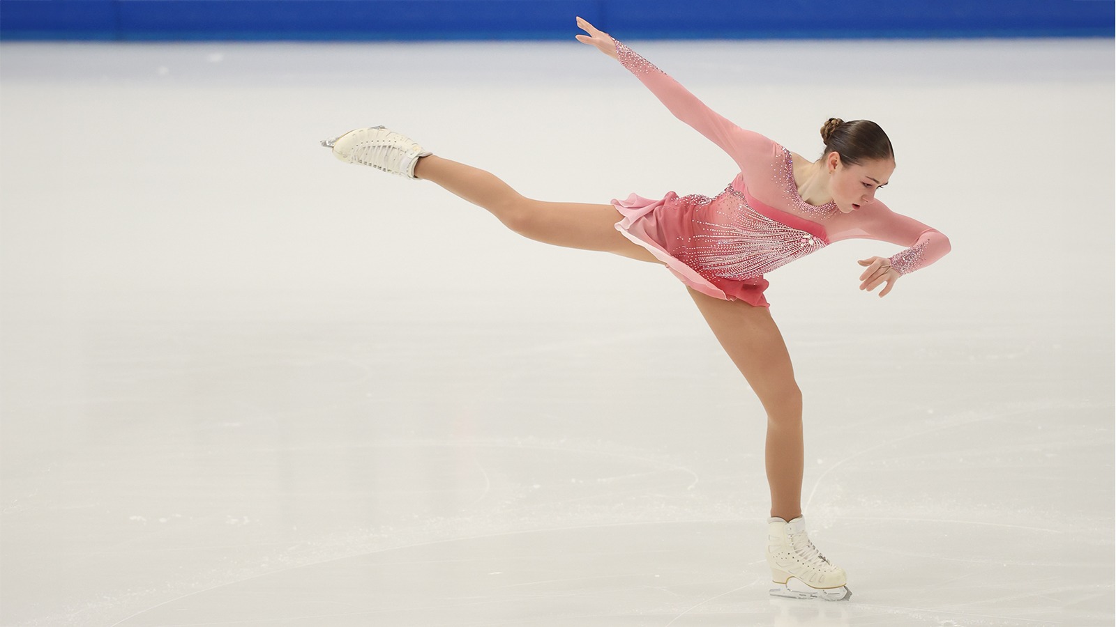 Sophie Joline von Felten gets ready to jump with one leg and one arm extended outward, and the other arm tucked in. Her blonde hair is put up in a bun and she is wearing a light pink long sleeve skating costume with jewel detailing down the front and on the sleeves.