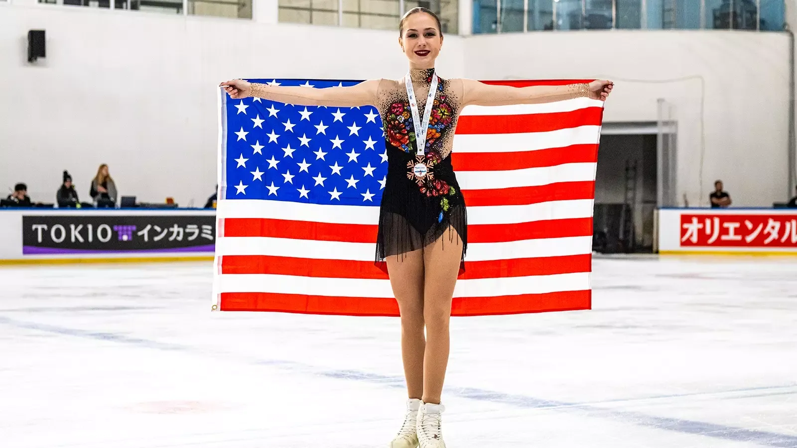 Sophie Joline von Felten stands proudly, smiling while holding a United States flag behind her with a bronze medal with a white ribbon around her neck. Sophie Joline is a young woman with blonde hair tied back in a bun wearing a black skating costume with multicolored flower detailing down the front and sheer long sleeves. 