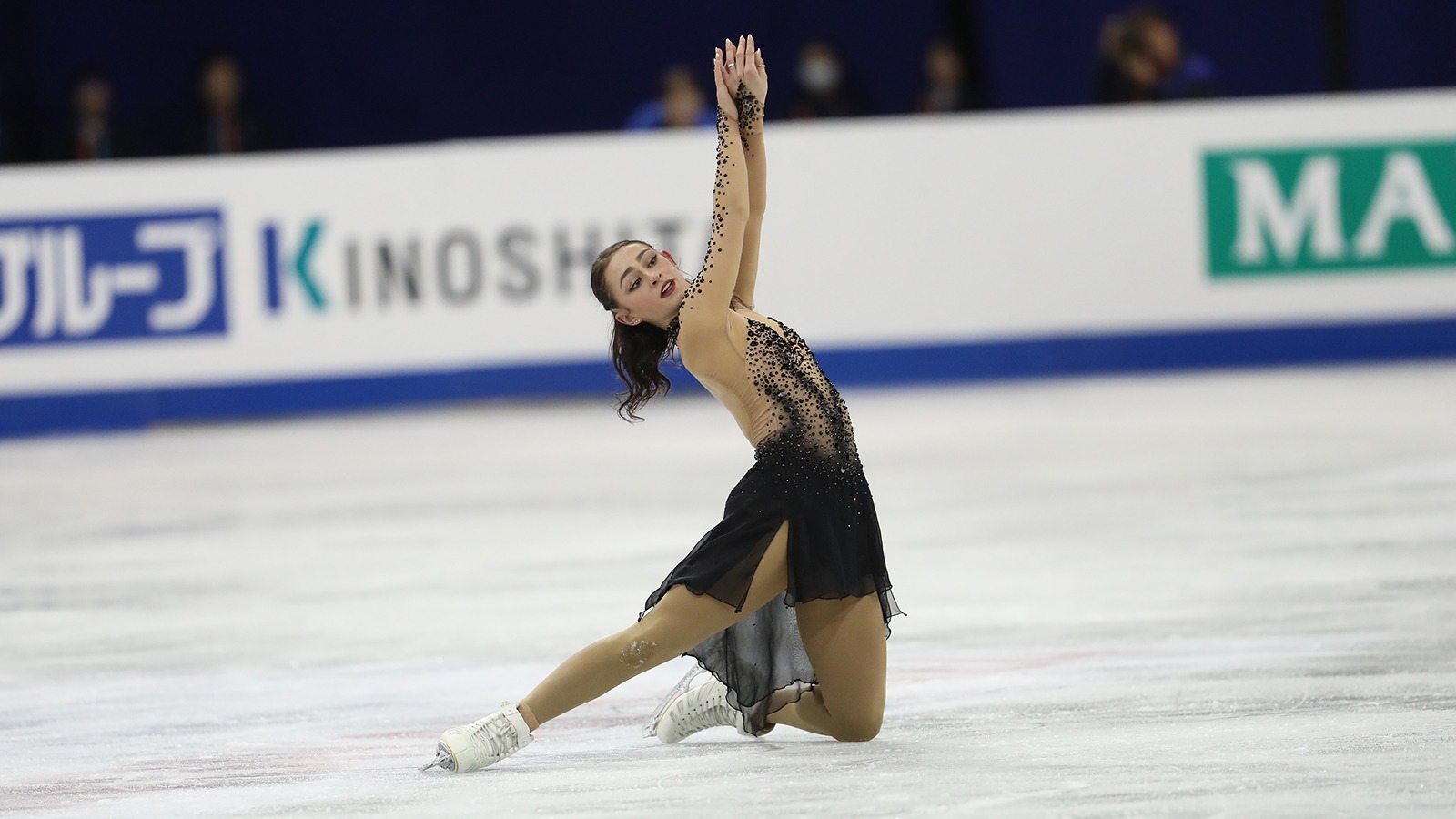 Ava Ziegler kneels on the ice with both arms raised above her head. She is a young woman with long brown hair wearing a black and tan skating costume. 