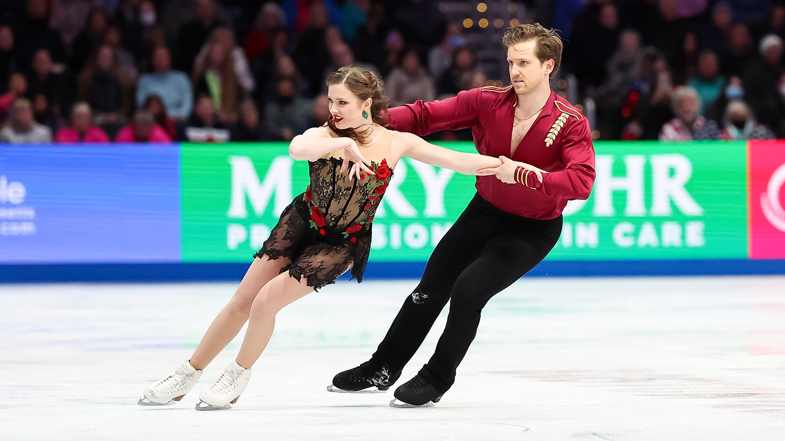 Christina Carreira and Anthony Ponomarenko skate together with Anthony skating behind Christina, holding on to her outstretched arm. Anthony is a young man wearing a long sleeve red top and Christina is a young woman with long brown hair wearing a black lace skating costume with red rose details. 