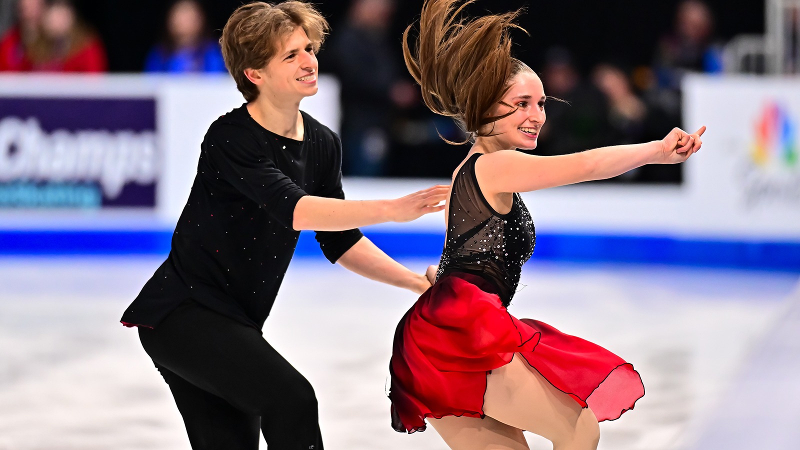 Oona Brown, dressed in a black top with a flowing red dress, and Gage Brown, dressed in all black, perform a fun routine at the 2025 U.S. Championships. Both have big smiles and a fun attitude.
